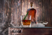 A Square Glass Of Whiskey With Ice And A Sprig Of Rosemary, And A Full Bottle Of Whiskey, Wrapped In Clouds Of Smoke, Stand On A Tray Against An Old Wooden Wall.
