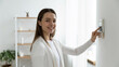 © fizkes - Happy young female client looking at camera, pressing buttons on smart house system. Smiling millennial woman using indoors temperature controller or air conditioning, setting alarm password.
