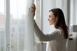 © fizkes - Side view happy attractive woman opening curtains, admiring cityscape at home or hotel room. Smiling millennial girl enjoying morning leisure weekend time or new day start, planning future indoors.