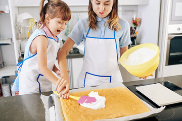 Wall Mural - Young mother in apron helping clumsy daughter to put cream frosting on baked cake