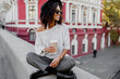 Street fashion look. Stylish black girl sitting on the bridge and holding cup of coffee or tea during her free time. Freelance woman. Wearing black hat and sunglasses.