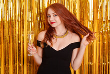 Happiness Woman Holding Glass Of Champagne, Touching Her Red Beautiful Hair, Posing Against Golden Tinsel On Background, Lady In Black Dress On Party.