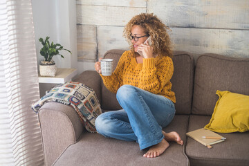 Wall Mural - Beautiful middle age woman with yellow sweater do phone call at home sitting on the couch and drinking coffee - relax lifestyle day for cheerful people - independence life concept