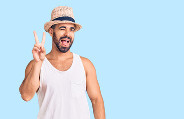 Young hispanic man wearing casual summer hat smiling with happy face winking at the camera doing victory sign. number two.