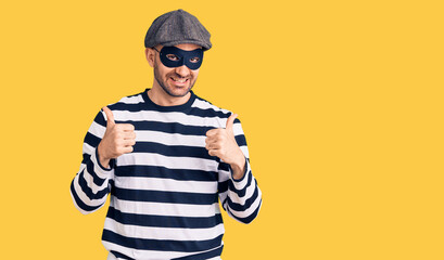 Young handsome man wearing burglar mask success sign doing positive gesture with hand, thumbs up smiling and happy. cheerful expression and winner gesture.