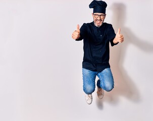 Middle age handsome hispanic man wearing glasses and cook uniform smiling happy. Jumping with smile on face doing ok sign with thumbs up over isolated white background