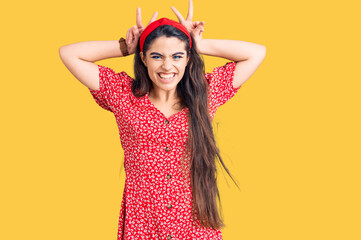 Brunette teenager girl wearing summer dress posing funny and crazy with fingers on head as bunny ears, smiling cheerful