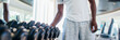 © twinsterphoto - African American man picking up a dumbbell from the rack and looking outside at gym. Male weight training person holding sport equipment with serous look in fitness center