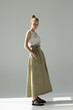 © gromovataya - Young woman posing in studio on a light background. Long green skirt, white summer T-shirt, black sandals. She stands sideways, hands in pockets.