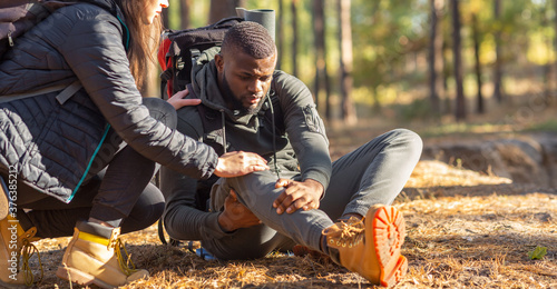 Unrecognizable woman comforting injured black guy, backpacking together by forest