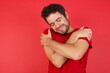 © Roquillo - Young handsome caucasian man wearing t-shirt over isolated red background Hugging oneself happy and positive, smiling confident. Self love and self care