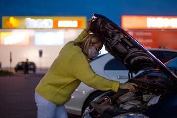 Wall Mural - Young Woman with coronavirus mask Looking In The Car Trunk in the car parking at night with bokeh background. women the car is broken road. Open the bonnet, check engine. Young woman reparing the car