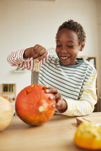 Vertical Medium Portrait Of Joyful Young Boy Carving Pumpkin With Kitchen Knife While Preparing For Halloween