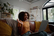 © StratfordProductions - Smiling young african american woman relaxing on the couch waving hand while talking to friends on a video call at home - young blogger working from home