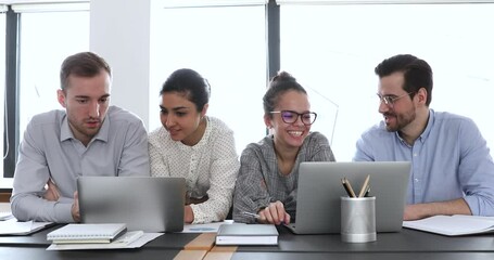 Canvas Print - Young multicultural workers group talking, looking at laptops and analyzing digital data sitting at workplace desk. Professional colleagues team discuss online project use devices in corporate office.