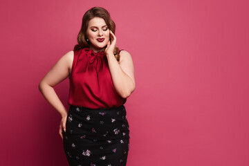 Confused plus-size model girl with bright makeup and with stylish hairstyle in a red blouse and skirt isolated at the pink background