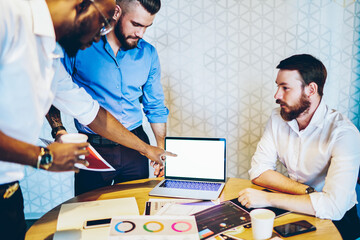 Multicultural male colleagues in formal wear talking during working process in office,successful proud ceo with dark skin pointing on mockup laptop computer with blank screen area for internet content