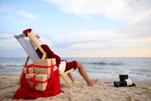 Santa Claus With Bag Of Presents Relaxing In Chair On Beach, Space For Text. Christmas Vacation