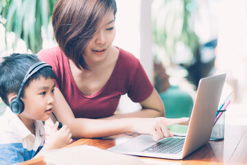 Wall Mural - Young asian mother and son using laptop computer for study and learning together at home, boy wearing headphone for e-learning with distancing, teacher or mom support child, education concept.