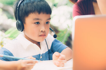 Wall Mural - Young asian mother and son using laptop computer for study and learning together at home, boy writing on notebook for homework and wearing headphone, teacher or mom support child, education concept.