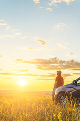 A young man in a shirt enjoys the sunset. A man and his car in a field against the backdrop of a brightly lit sunset sky. Local travel concept. Vertical orientation, space for text.