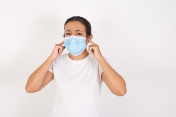 Young arab woman wearing medical mask standing over isolated white background covering ears with fingers with annoyed expression for the noise of loud music. Deaf concept.