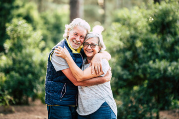 Beautiful elderly lifestyle senior couple hug and stay in love touching during outdoor leisure activity in the wood forest - green nature background and happy old people together