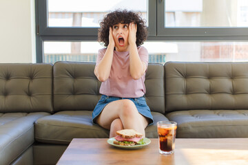 Wall Mural - young pretty arab woman having a sandwich sitting on a sofa at home