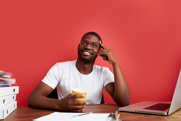 portrait of smiling african man sitting at office desk, drinking cup of coffee in the morning, isolated over red background