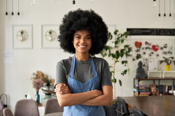 Happy African American young woman with Afro hair modern cafe small business owner, female waitress in reopened restaurant looking at camera standing arms crossed in cozy cafe interior. Portrait.