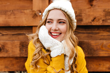 Wall Mural - Portrait of a smiling girl in cute warm clothes touching her face and curly hair and looking to the left