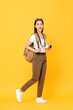 Full length portrait of smiling cute young Asian woman tourist with camera waling on isolated yellow studio background