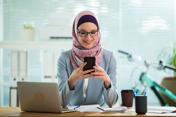 Wall Mural - Beautiful young working woman in hijab and suit sitting in office, using smart phone. Portrait of confident muslim businesswoman. Modern light office with big window. 