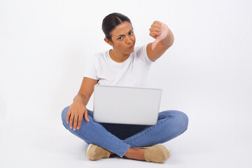 Sticker - Discontent European woman shows disapproval sign, keeps thumb down, expresses dislike, frowns face in discontent, dressed in white shirt, isolated over gray background. Body language concept.