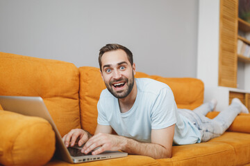 Wall Mural - Shocked amazed surprised attractive young bearded man 20s wearing casual blue t-shirt working on laptop pc computer lying on couch resting relaxing spending time in living room at home.