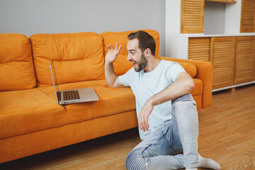 Canvas Print - Excited surprised young bearded man 20s in casual blue t-shirt working on laptop pc computer making video call greeting with hand sitting at floor near couch spending time in living room at home.