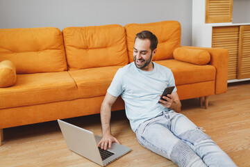 Canvas Print - Cheerful young bearded man 20s in casual blue t-shirt working on laptop pc computer using mobile cell phone typing sms message sitting at floor near couch resting spending time in living room at home.