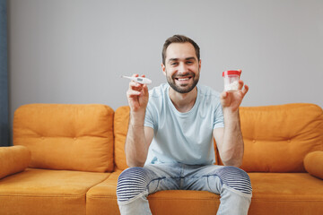 Canvas Print - Smiling cheerful attractive young bearded man in casual blue t-shirt hold medication tablets aspirin pills and thermometer looking camera sitting on couch spending time in living room at home.