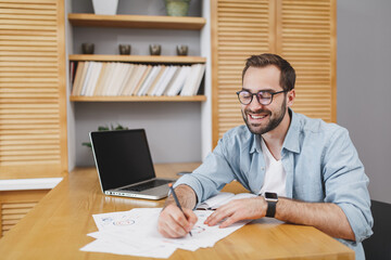 Canvas Print - Cheerful smiling attractive young bearded business man 20s wearing blue shirt glasses sitting at desk working on laptop pc computer writing on papers document at home or office.