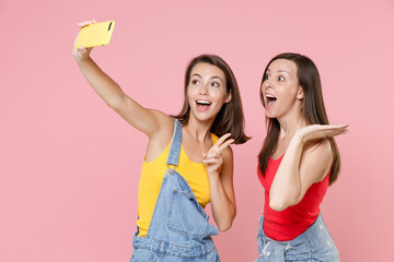 Wall Mural - Two surprised funny young brunette women friends 20s wearing casual denim clothes doing selfie shot on mobile phone showing victory sign isolated on pastel pink colour background, studio portrait.