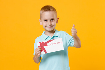 Wall Mural - Smiling cute little fun male kid boy 5-6 years old wearing stylish blue turquoise t-shirt polo hold in hand mockups of gift certificate isolated on yellow color wall background, child studio portrait.