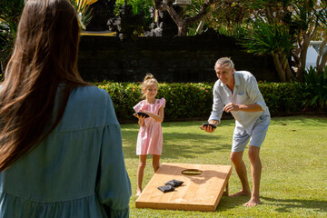 Happy family playing cornhole game outdoor on sunny summer day. Parents and children playing bean bag toss