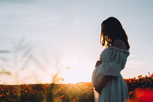 Silhouette Of Beautiful Pregnant Woman Standing On Poppies Field In Sunset. Pregnant Woman In White Dress Relaxing Outdoors. Copy Space.