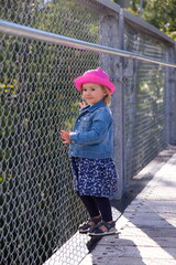 Wall Mural - Vertical view of cute blonde toddler girl in jeans jacket, blue dress and pink straw hat standing on walkway leaning against wire mesh railing with unsure expression, Quebec City, Quebec, Canada