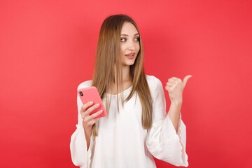 Eastern European woman holding bucket with popcorn standing over isolated red background using and texting with smartphone  pointing and showing with thumb up to the side with happy face smiling