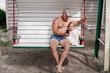 Athletic man and a three year old girl on a white swing near the house. Father shows his daughter's phone. Dad and baby. Recreation in the yard