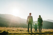 © Serhii - Family with kid hiking in mountains. Young tourists on top of a mountain enjoying valley view sunset. Happy mom, dad with a backpack and daughter. Holiday trip concept. World Tourism Day. Back view.
