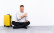 © luismolinero - Young handsome man sitting on the floor with a suitcase pointing finger to the side