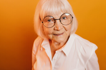 Top view of old smiling woman with gray hair and gray eyes, wearing round glasses and white clothes. Beautiful woman isolated over bright orange background. Looking at the camera.
