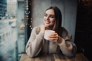 Wall Mural - Prosperous hipster girl 20s with perfect veneers laughing during resting time in cafe interior, sincerely female customer with tea cup for warming smiling during cafeteria recreation on weekend